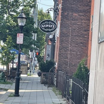 Gypsys - Sign outside - The image shows a city sidewalk adjacent to a brick building with a hanging sign for Gypsy's. The sidewalk has street lamps, trees, and outdoor seating with chairs and tables. Utility poles and wires are visible in the background, along with other buildings and street signs. The scene appears to be in a downtown or commercial area with pedestrian amenities. - Dive Bars Near Me - Covington, Kentucky