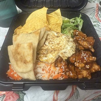 Fusion Crunch - Chimi BBQ. Comes with pita bread, rice, tortilla chips and fried egg - A black plastic takeout container contains a meal with white rice topped with shredded carrots, grilled chicken pieces coated in sauce, a fried egg, tortilla chips, and folded tortillas. A small side salad with lettuce is also included. The container is placed on a table with a patterned tablecloth. - Burgers Near Me - Bloomington, Illinois