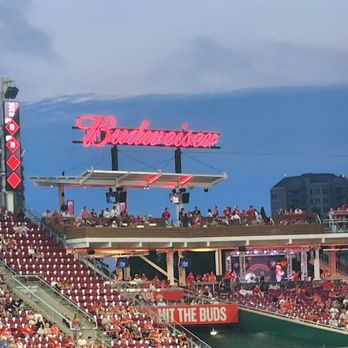 Cincinnati Reds - The image shows a baseball stadium with a large crowd of spectators seated in the stands. A bright Budweiser sign is visible above the seating area, and a large screen displays game footage. The sky is partly cloudy, and the stadium features multiple levels of seating and advertising banners. - Professional Sports Teams Near Me - Cincinnati, Ohio