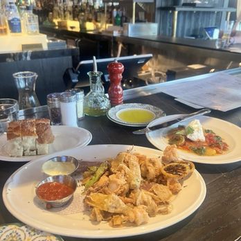 Valentina's - The image shows a restaurant table with a plate of fried calamari accompanied by two dipping sauces, a piece of bread, and a salad with cheese and vegetables. The background includes a bar area with bottles, glasses hanging upside down, and a window with natural light. The table has condiments such as olive oil, salt, pepper, and a red pepper grinder. - Italian Near Me - Dublin, Ohio