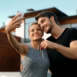 Haskell Real Estate - Young couple taking a photo with their phones in front of their new home.
