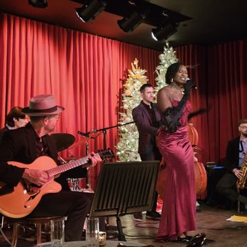 Monarque - Live band and jazz singer who also happens to be an army veteran. I love that! - A live jazz band performs on stage at Monarque in Baltimore, MD, with a female singer in a red dress holding a microphone. The band includes a guitarist, a saxophonist, and other musicians, with Christmas trees and red curtains as the backdrop. The scene captures a musical performance in a venue with stage lighting. - French Near Me - Baltimore, Maryland