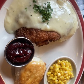 99 Restaurants - Chicken dinner with mashed potatoes and corn - The image shows a plate with a serving of chicken fried steak covered in white gravy and garnished with chopped herbs. Accompanying the main dish are mashed potatoes, a small bowl of cranberry sauce, and a side of cooked corn kernels. The meal is presented on a white plate with a red rim, placed on a dark wooden table. - American Near Me - Wareham, Massachusetts