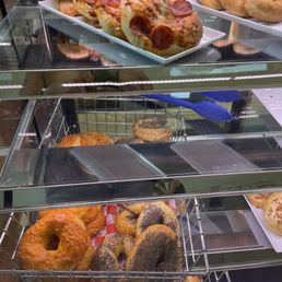 The image shows a glass display case with multiple shelves containing baked goods. The top shelf features pepperoni pizza bagels on a white rectangular tray. The lower shelves hold assorted bagels, including plain and poppy seed varieties, arranged on metal racks. The case is situated in a bakery or cafe setting, with some equipment and a person partially visible in the background.