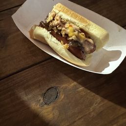 Diddy Dog - A hot dog with toppings including chopped onions and sauce is presented in a bun on a white paper tray. The tray is placed on a wooden table, and a crumpled napkin is visible in the background. The image is taken from an angle showing the side of the hot dog.