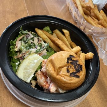 The Den Cape Cod - lobster roll app - The image shows a seafood meal served in a black plastic container, featuring a lobster roll with a toasted bun, French fries, and a side salad with lettuce, cucumbers, and red onions. A lemon wedge is also included. In the background, there is a clear plastic takeout box filled with more French fries. The setting appears to be a casual dining or takeout environment. - American Near Me - Dennis Port, Massachusetts