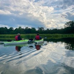 A Canoe To You - Two people from rear kayaking in wide tree-edged creek.