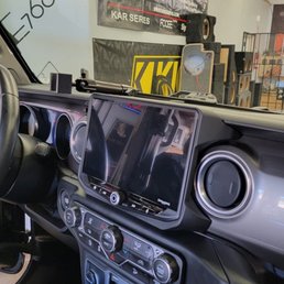 Stinger Heigh10 multimedia display. - The image depicts the dashboard of a vehicle with a large central touchscreen, circular air vents, and various control buttons, inside a workshop with audio equipment in the background.