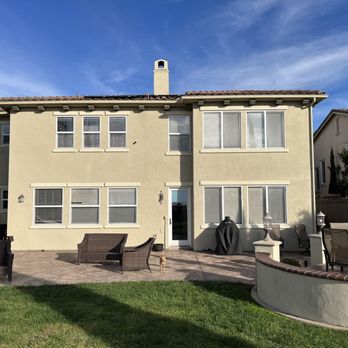 Appleton Design & Construction - Before - The image shows the rear view of a two-story residential house with a beige stucco exterior and a tiled roof. The backyard includes a paved patio area with outdoor seating, a grill covered with a black cover, and a curved low wall with decorative lamps. A grassy lawn extends in the foreground, and the sky above is clear with some clouds. The scene suggests a well-maintained outdoor space for leisure and gatherings. - General Contractors Near Me