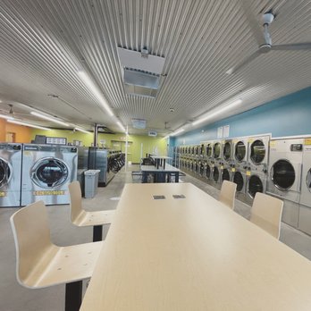City Laundry - Phoenix - The lounge countertop with plug-ins - The image shows the interior of a laundromat with multiple washing machines and dryers arranged along the walls. The ceiling has a metallic finish with fluorescent lighting. A large wooden table with chairs is positioned in the foreground, and the space includes a trash bin and a few small tables in the background. The walls are painted in different colors, including blue and green, creating a vibrant atmosphere. - Laundromat Near Me - Phoenix, Arizona
