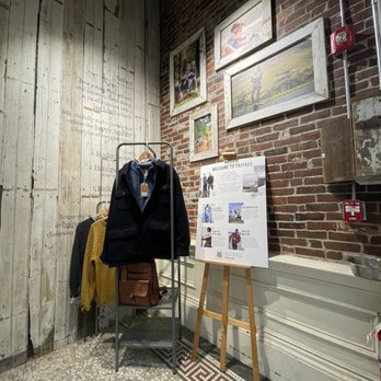 FatFace - Interior of a clothing store featuring jackets on a rack, framed photos on brick walls, and a welcome sign on an easel. - Women's Clothing Near Me - Portland, Maine