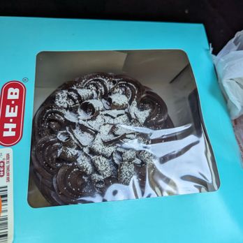 H-E-B - Chocolate cake on its way to birthday consumption - A chocolate cake with decorative swirls and powdered sugar on top is visible inside a blue H-E-B bakery box with a clear plastic window. The cake appears to be a dessert item, possibly for a celebration, and the box is placed on a dark surface with a receipt partially visible underneath. - Grocery Near Me - Mckinney, Texas