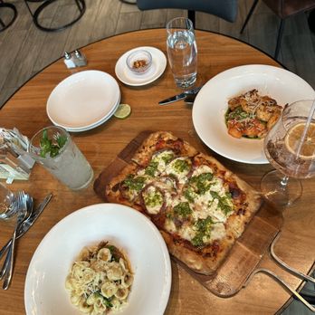 the Albert - The image shows a round wooden table set with a variety of dishes and drinks at a restaurant. There is a rectangular pizza with cheese, tomato, and green pesto, a bowl of pasta with cheese and vegetables, and two beverages—one with a lemon slice and a straw, and a glass of water. Plates, utensils, a small bowl of chili flakes, and a lime wedge are also visible on the table. The background includes chairs and part of the restaurant interior. - New American Near Me - Chicago, Illinois