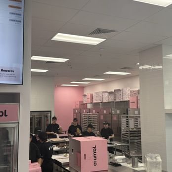 Crumbl - La Crosse - Inside of Crumbl (La Crosse, WI) - The image shows the interior of a Crumbl bakery kitchen with several employees dressed in black uniforms and caps. They are working at counters with baking trays and equipment. The background features pink walls, stacked boxes, and stainless steel racks. The setting appears clean and organized, with a modern, minimalistic design. - Bakeries Near Me - Onalaska, Wisconsin