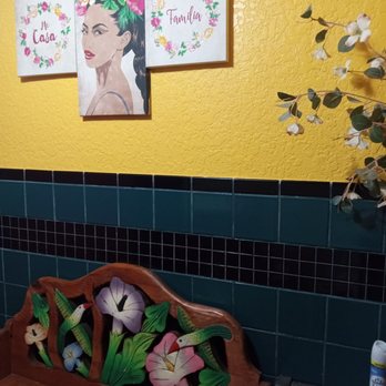 El Chef - Pretty restroom - The image shows three pieces of decorative wall art hung on a yellow textured wall. The artwork includes floral designs and the words "Mi Casa" and "Familia," along with a portrait of a woman with a floral crown. Below, a wooden bench with carved floral and bird motifs is positioned against a green tiled wall with black accents. A roll of paper towels and a spray can are placed on a small surface next to the bench, and a branch with white flowers extends into the frame from the right side. - Mexican Near Me - Douglas, Arizona