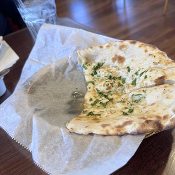 Namaste - Garlic Naan....light and luscious tasting with every bite! - A piece of garlic naan bread, garnished with chopped herbs, is in a basket lined with parchment paper on a wooden restaurant table. A glass of water with a straw is visible in the background, and the restaurant interior features wooden flooring and chairs. - Indian Near Me - Rio Rancho, New Mexico