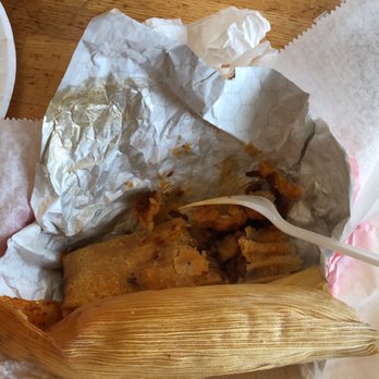 Gigio's Pizzeria - Tamale - The image shows a partially eaten tamale wrapped in a corn husk, placed on a paper-lined tray. The tamale appears to have a filling that has been partially consumed, with some crumbs and remnants visible. The tray is on a wooden table, with a plastic fork inserted into the tamale. In the background, there is a cup with a lid and straw, and some other food items. The caption indicates the tamale is from a Midwest pizzeria that also sells gyros. - Pizza Near Me - Evanston, Illinois