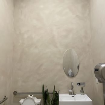 Kora - Single very large bathroom - The image shows a small bathroom with beige, textured walls. There is a white toilet with the lid open, a small rectangular white sink with a faucet, and a tall green plant placed between the toilet and sink. A small oval mirror is mounted on the wall above the sink. Metal handrails are installed on the walls for support, and a wall-mounted soap dispenser is visible on the right side. The space appears clean and minimalistic. - Filipino Near Me - Sunnyside, New York