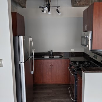 VRC Management - Kitchen Before - The image shows a kitchen with a stainless steel refrigerator on the left, a black countertop with a sink, and wooden cabinets above and below. A microwave is mounted above the stove, which is also stainless steel. The ceiling has an exposed concrete beam and a modern track lighting fixture. The walls are painted light gray, and the flooring is dark wood or laminate. - General Contractors Near Me - Chicago, Illinois