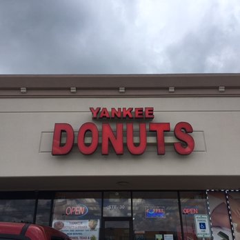 Yankee Donuts & Drinks - sign to look for - The image depicts the storefront of Yankee Donuts & Drinks in Spring, TX, with a prominent sign displaying the business name in red letters. The building has a beige facade with large glass windows and doors, some of which have illuminated signs indicating the store is open. The sky above is overcast with dark clouds, and part of a vehicle is visible in the lower left corner. - Donuts Near Me - Spring, Texas
