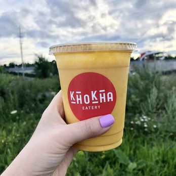 Khokha Eatery - Mango Milkshake (lassi) - A mango lassi in a clear plastic cup featuring a red Khokha Eatery logo is held by a person with light-colored skin and purple nail polish. The background shows a partly cloudy sky and green vegetation, indicating an outdoor setting. The image emphasizes the beverage and branding of Khokha Eatery, a restaurant offering Indian cuisine. - Pakistani Near Me - Ottawa, Ontario