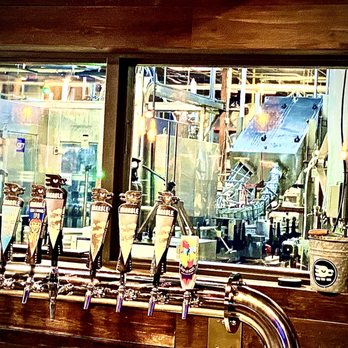 Marble Brewery - The image shows a row of beer taps mounted on a wooden bar counter in a brewery taproom. Behind the taps, a window provides a view into the brewing area, where brewing tanks, pipes, and scaffolding are visible. The scene includes some small items like a sanitizer bottle and a bucket on the window ledge. The setting suggests an indoor space dedicated to craft beer serving and brewing activities. - Breweries Near Me - Albuquerque, New Mexico