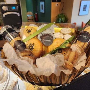 Masuda’s Cafe - A gift basket from Masuda's Cafe features an assortment of cookies, including some with black sesame seeds, and small jars labeled with the cafe's branding. The basket is lined with tissue paper and decorated with green ribbons, presenting a selection of baked goods and treats suitable for gifting or sampling. The background shows a cafe interior with framed pictures and a green wall. - Cafes Near Me - Amherst, Massachusetts