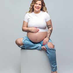 Kimberly Ogden Portraits - Pregnant woman in white crop top and ripped jeans sitting on white cube, showing baby bump with tattoo on arm.