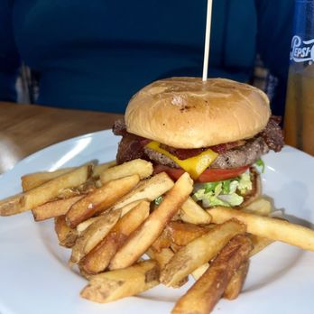 Bearded Monkey - A burger with a bun, lettuce, tomato, cheese, beef patty, bacon, and a skewer on top is served with a side of French fries on a white plate. A glass of soda with ice and a Pepsi logo is visible in the background, along with a person wearing a blue shirt. The setting appears to be a casual dining environment. - Chicken Wings Near Me - Fredericksburg, Virginia