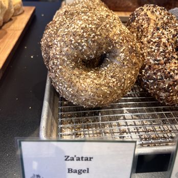 Cellar Door Cafe - Za'atar bagel - A close-up of a Za'atar bagel topped with sesame seeds and herbs, resting on a wire cooling rack at a bakery. The bagel has a round shape with a hole in the center, and the surface is coated with seeds and herbs. A label in front of the bagel reads 'Za'atar Bagel,' indicating the type of baked good. The background shows other baked items and a bakery display setting. - American Near Me - Lawrence, Kansas