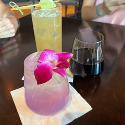 Three cocktails are on a dark wooden table: a pink drink with an orchid garnish, a yellow drink with lime and a straw, and a dark red drink in a wine glass.