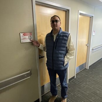 The Pit - A man with glasses, dressed in a beige shirt and a blue puffer vest, stands in a hallway with beige walls and dark flooring. He is pointing at a door with a sign indicating 'Lobo Basketball.' The hallway has two closed wooden doors with metallic frames and a handrail on the wall. - Stadiums & Arenas Near Me - Albuquerque, New Mexico