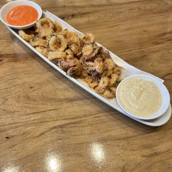 Prato - Pretzel crusted calamari. Very good. Couldn't taste any pretzel crust though. With Salsa Rossa and Grain Mustard Aioli. - A serving of fried calamari rings, garnished with chopped green herbs, is arranged on a long, narrow white platter. The dish includes two small bowls of dipping sauces: one orange-colored and the other creamy with herbs. The platter is placed on a wooden table, and the presentation emphasizes the appetizer's crispy texture and variety of sauces. - Pizza Near Me - Winter Park, Florida