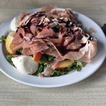 The Local - Peaches & Cream  THIS SALAD IS THE BEST!! - A salad featuring mixed greens, sliced peaches, prosciutto, and dollops of cheese, topped with a drizzle of balsamic glaze, presented on a white plate placed on a wooden surface. The ingredients are arranged to showcase the combination of fresh fruit, cured meat, and cheese, with the glaze adding a finishing touch. - Cocktail Bars Near Me - Babylon, New York