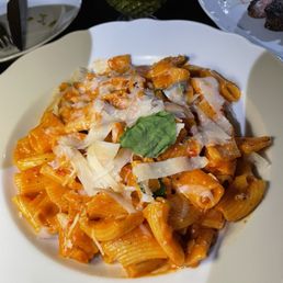 Spicy Vodka Rigatoni - A bowl of rigatoni pasta with tomato-based sauce, garnished with cheese shavings and a basil leaf, served at a restaurant.