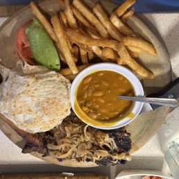 Ecuadorian cuisine - A meal plate includes French fries, a fried egg, a slice of tomato, an avocado, a grilled meat portion topped with sautéed mushrooms, a small bowl of beans, and a fried plantain. The food is arranged on a wooden platter, with a spoon in the bean bowl. The setting appears to be a casual dining environment, emphasizing traditional South American dishes.