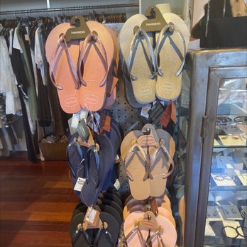 Jaffi's - A retail store features a display of Havaianas flip-flops in multiple colors, including pink, yellow, blue, and black, hanging on a pegboard. The flip-flops are arranged in pairs with tags attached. In the background, clothing racks with various garments and a jewelry display case with accessories are visible. The store has a wooden floor and appears to sell clothing, accessories, and footwear. - Women's Clothing Near Me - Neptune Beach, Florida