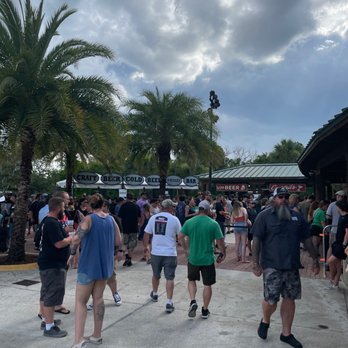 St Augustine Amphitheatre - Pretty good crowd already for the Flogging Molly show. - The image shows a crowd of people standing and walking outside a bar area at an outdoor event. Palm trees are visible, and the sky is partly cloudy. The scene includes signage indicating craft beer, cold beer, and a bar, with some individuals wearing casual clothing and sunglasses. The setting appears to be in a warm climate, possibly during late afternoon or early evening. - Music Venues Near Me - Saint Augustine, Florida