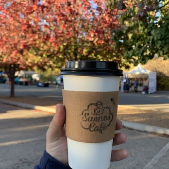 Sugar N Glitz - Hot chocolate - A disposable coffee cup with a black lid and a cardboard sleeve reading 'Sueño Café' is held in front of a parking lot with trees and tents. - Bakeries Near Me