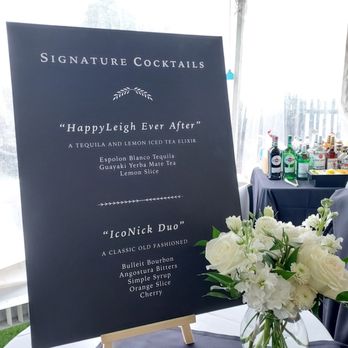 Creative Catering - A black menu board displays signature cocktails and a floral arrangement with white flowers on a table at an event. - Caterers Near Me - Beverly, Massachusetts