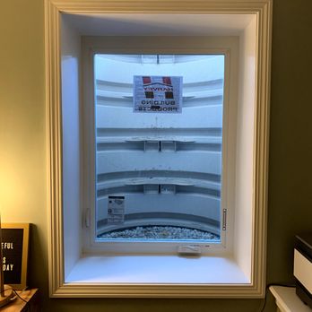 LB Special Projects - Egress window - The image shows a newly installed egress window in a residential room, with a view of a balcony outside. The window frame is white, and the interior wall around it is painted green. The window sill is clear, and there is a power outlet below the window. Outside, the balcony has curved railings and a sign indicating construction work. The ceiling has decorative molding, and part of a ceiling panel is visible at the top of the image. - General Contractors Near Me - Middleton, Massachusetts
