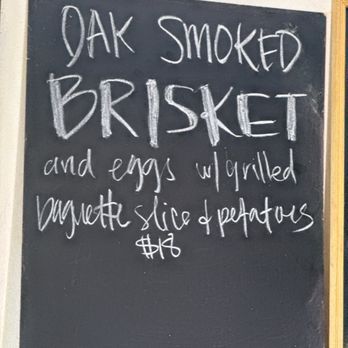 ACME Cafe - Saturday Special - A black chalkboard sign features handwritten white chalk text advertising oak smoked brisket, served with grilled baguette slices and potatoes, priced at $15. The sign is positioned against a white wall with a wooden frame visible on the right side. - New American Near Me - Salem, Oregon