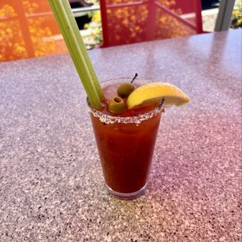 Egg N' Joe - Bloody Mary - A Bloody Mary cocktail with a salted rim, garnished with green olives, a lemon wedge, and a celery stalk, placed on a speckled table. - American Near Me - Gilbert, Arizona