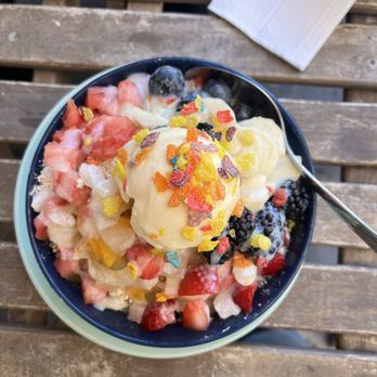 Cafe BT - An aerial view of the shaved ice - A bowl of shaved ice dessert features a scoop of vanilla ice cream topped with colorful sprinkles, surrounded by chopped strawberries, blueberries, and other fruit pieces. The dessert is placed on a wooden outdoor table, with a spoon inserted into the bowl and a paper napkin nearby. The setting appears casual and outdoor, suitable for enjoying a cold treat. - Coffee & Tea Near Me - Walnut, California