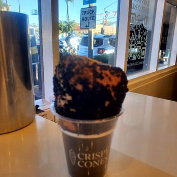 Crispy Cones - A chocolate ice cream cone is served in a clear plastic cup with the Crispy Cones logo printed on it. The ice cream appears to be chocolate-flavored with a textured surface. The cup is placed on a light-colored cafe table near a window, through which outdoor parking and street signs are visible, including a parking sign and a truck route sign. The scene is illuminated by natural daylight. - Desserts Near Me - Carlsbad, California