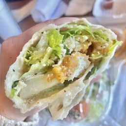 Chicken Wrap - A crispy chicken wrap filled with lettuce, cheese, and caesar dressing, held in a person's hand, with a casual dining setting in the background.