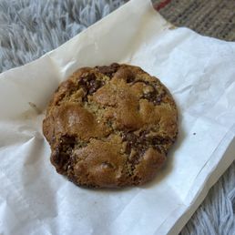 Chocolate Chip Cookie - A chocolate chip cookie with a golden-brown crust and visible chocolate chunks is placed on white parchment paper. The background includes a soft, furry surface and a textured woven fabric, suggesting a cozy indoor setting. The cookie appears freshly baked and slightly cracked on top.