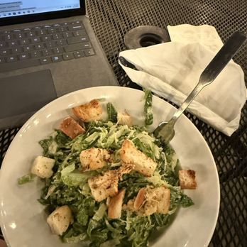 Willy McCoy's - Ceased salad - A Caesar salad with croutons and lettuce is on a white plate, placed on a black metal table near a laptop and a napkin with utensils. - American Near Me - Champlin, Minnesota