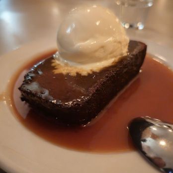 The Snail - Sticky toffee pudding - A dessert plate holds a slice of rich, dark chocolate cake topped with a scoop of vanilla ice cream. The cake is drizzled with caramel sauce, which pools around the base. The dessert is presented on a white plate, with a metal spoon placed nearby. The background shows a blurred restaurant environment with glasses and tableware, indicating a dining setting. - American Near Me - Brooklyn, New York