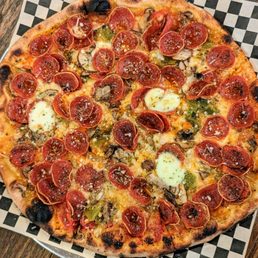 Lobo | Instagram: @telephonesmoothie - A round pizza with a golden-brown crust topped with pepperoni slices, melted cheese, green peppers, mushrooms, and dollops of cheese. The pizza is placed on a checkered paper-lined tray on a wooden table, showing a variety of toppings and a slightly charred crust.